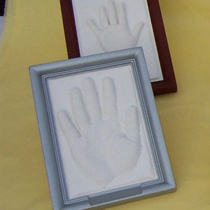 May include: Two framed handprints, one in a silver frame and one in a brown frame, on a yellow surface.