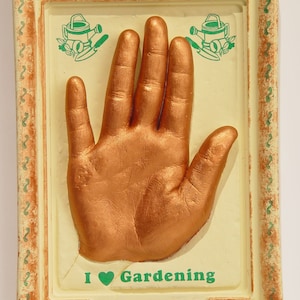 May include: A gold handprint in a white frame with a green border. The handprint is surrounded by a green and white design of watering cans and plants. The text "I ♥ Gardening" is below the handprint. A small plaque below the text reads "Rhonda Summer 2003".