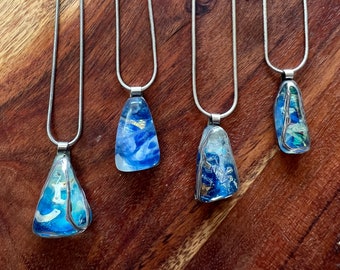 Puede incluir: Cuatro collares de cadena plateada con colgantes de vidrio azul y dorado. Los colgantes son de diferentes formas y tienen una variedad de patrones azules y dorados.