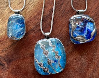Puede incluir: Tres collares de cadena plateada con colgantes de vidrio azul y plateado. Los colgantes son rectangulares y presentan una variedad de patrones azules y plateados.