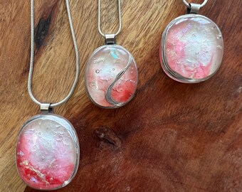 Puede incluir: Tres collares de cadena plateada con colgantes de forma ovalada. Cada colgante está relleno de resina rosa y blanca con un acabado brillante.