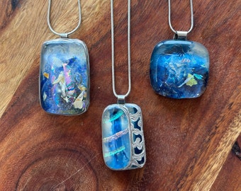 Puede incluir: Tres collares de cadena plateada con colgantes de vidrio azul. Los colgantes son rectangulares y presentan vidrio azul y verde iridiscente con detalles plateados.