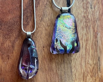 Puede incluir: Dos colgantes de vidrio iridiscente en cadenas plateadas. Los colgantes tienen forma de lágrima y presentan una gama de colores púrpura, azul y dorado.
