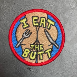 Könnte beinhalten: Gestickter Aufnäher mit einem hellbraunen Hintern-Design, einer silbernen Gabel und einem Messer sowie dem Text "I EAT THE BUTT" in Gelb. Der Aufnäher hat einen blauen Mittelteil, einen roten Rand und einen schwarzen Hintergrund.