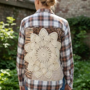 May include: A long-sleeve button-down shirt featuring a brown, white, and black plaid pattern. The back panel is embellished with a large, circular, cream-colored crocheted doily centered over mixed-texture fabric patches including animal prints.