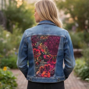 May include: A blue denim jacket featuring a large, rectangular back panel with a vibrant, multi-colored floral and paisley print in shades of magenta, orange, yellow, and black. The jacket has a classic collar, long sleeves, and buttoned cuffs.