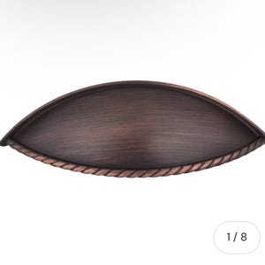May include: A bronze-colored cabinet pull with a curved, cup-like design. The pull has a textured surface and a rope-like detail along the bottom edge. The hardware is designed for easy installation and adds a touch of elegance to any cabinet or drawer.