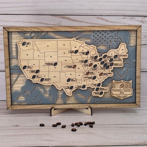May include: A wooden map of the United States with a gray and white background. The map is framed in a light brown wood frame and has a small sign that reads "Football Travel Map".