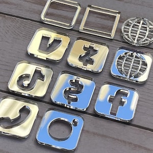 May include: A collection of shiny, silver-colored social media and communication icons. The set includes the Instagram, Facebook, TikTok, and phone icons, along with a globe and other geometric shapes. The icons are arranged on a wooden surface.