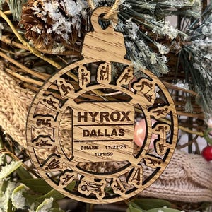 May include: Wooden Christmas ornament with the text "HYROX DALLAS" and the date "11/22/25". The ornament is circular with cut-out designs and a string for hanging. The background includes greenery and a woven basket.