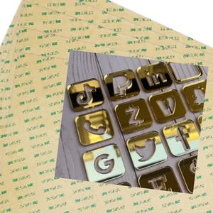 May include: Gold-colored social media icons, including Facebook, Twitter, Instagram, TikTok, LinkedIn, YouTube, Pinterest, and Snapchat, are arranged in a grid pattern on a wooden surface. The icons are made of acrylic and have a mirrored finish.