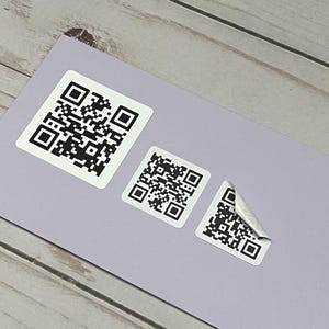 May include: Three white QR code stickers of varying sizes are displayed on a light purple surface. One corner of the smallest sticker is peeled back, revealing the adhesive. The background is a light wood grain.