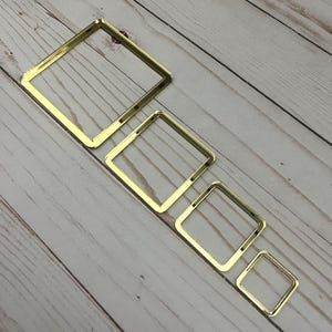 May include: Four gold-colored square frames of varying sizes arranged diagonally on a light wood-grain surface. The frames are hollow and have a reflective, metallic finish. The smallest frame is approximately 1 inch square.