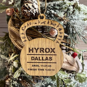 May include: Wooden ornament in the shape of a kettlebell, engraved with "HYROX DALLAS" and athlete's name and finish time. The ornament is decorated with small figures depicting workout exercises. It hangs from a natural twine string.