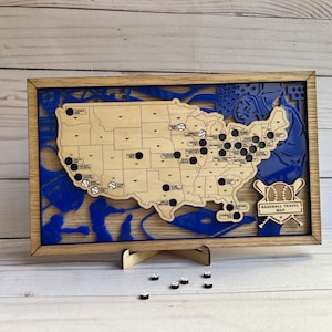 May include: A wooden map of the United States with blue and brown accents. The map is decorated with baseball-themed imagery and features black dots representing baseball stadiums. The text "Baseball Travel Map" is displayed at the bottom of the map.