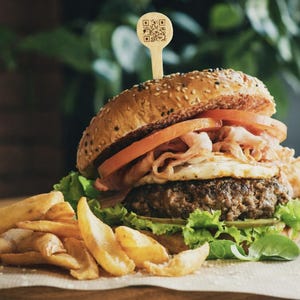 May include: A large burger with a sesame seed bun, tomato slices, bacon, and a fried egg. The burger sits on a bed of lettuce and is served with a side of golden french fries. A wooden pick with a QR code is in the burger.