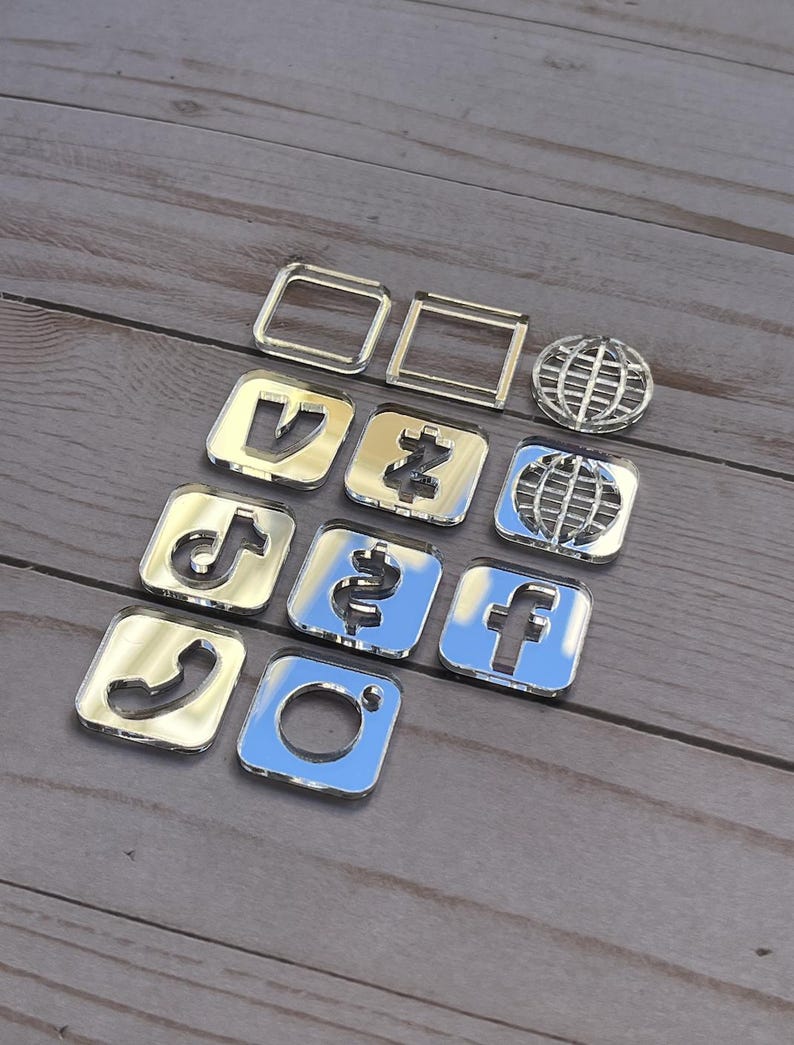 May include: A collection of shiny, silver-colored social media and communication icons. The set includes the Instagram, Facebook, TikTok, and phone icons, along with a globe and other geometric shapes. The icons are arranged on a wooden surface.