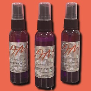May include: Three purple body spray bottles with clear caps and black spray nozzles. Each bottle has a label with the text "OM", "2oz Body Spray", and "SHAKE BEFORE USE". The background is a solid orange color.