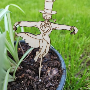 May include: A wooden cutout of a whimsical character with a top hat, bow tie, and a pipe. The character is standing in a pot of soil with green grass in the background.