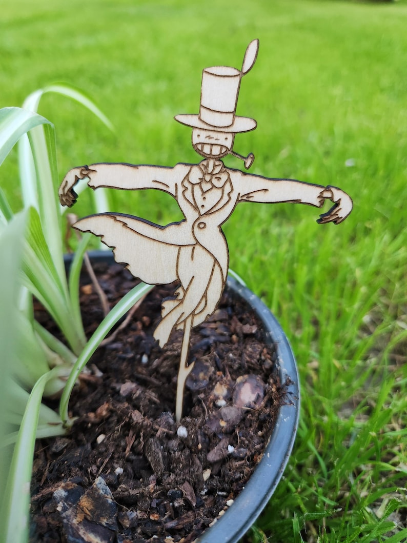 May include: A wooden cutout of a whimsical character with a top hat, bow tie, and a pipe. The character is standing in a pot of soil with green grass in the background.