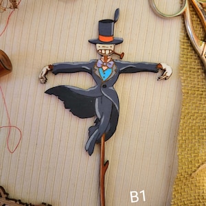 May include: A wooden cutout of a scarecrow wearing a black top hat with an orange band, a gray suit, a blue bow tie, and a pipe. The scarecrow is holding a stick with a branch at the top. The cutout is labeled "B1".