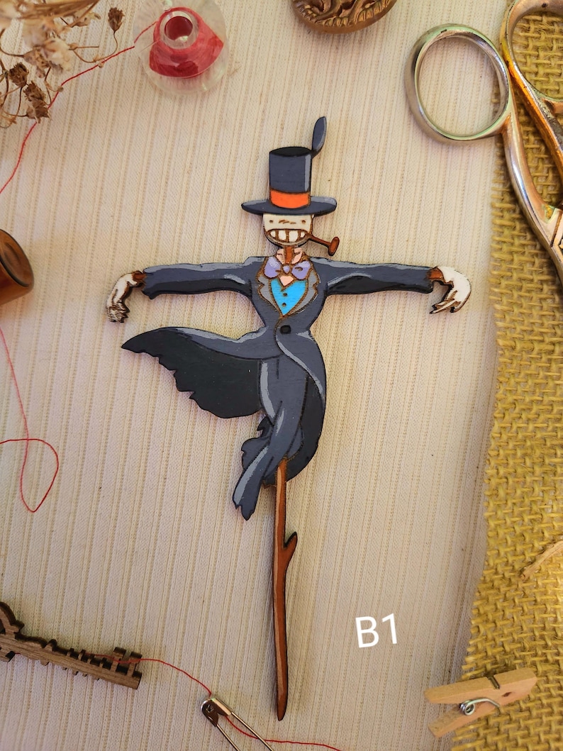 May include: A wooden cutout of a scarecrow wearing a black top hat with an orange band, a gray suit, a blue bow tie, and a pipe. The scarecrow is holding a stick with a branch at the top. The cutout is labeled "B1".