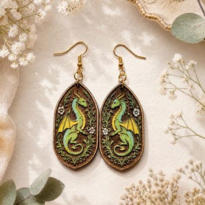 May include: A pair of oval-shaped wooden drop earrings featuring a detailed illustration of a green dragon with yellow wings, surrounded by intricate floral vines. The earrings are suspended from gold-toned metal hooks and have a dark wood finish.