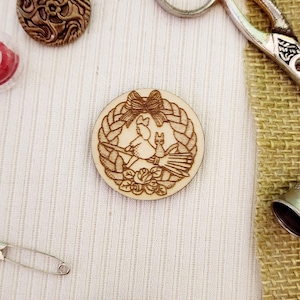 May include: A wooden circle with a carved design of a cat and a bird in a wreath with a bow. The circle is on a white fabric background.