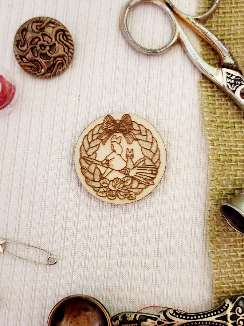 May include: A wooden circle with a carved design of a cat and a bird in a wreath with a bow. The circle is on a white fabric background.