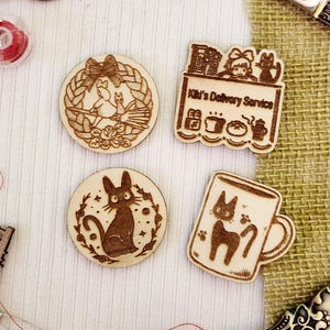 May include: Four wooden laser-cut ornaments featuring characters from the animated film Kiki's Delivery Service. The ornaments depict Kiki on a broomstick, Kiki's cat Jiji, Kiki's delivery service, and a mug with Jiji on it.