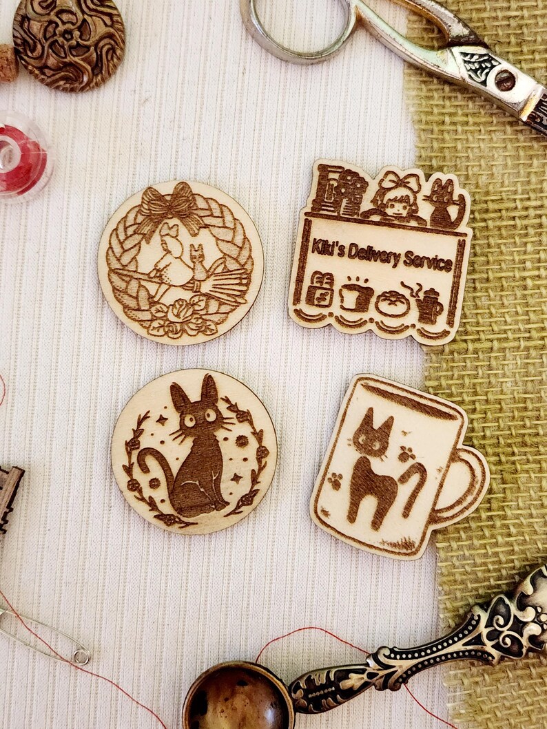 May include: Four wooden laser-cut ornaments featuring characters from the animated film Kiki's Delivery Service. The ornaments depict Kiki on a broomstick, Kiki's cat Jiji, Kiki's delivery service, and a mug with Jiji on it.