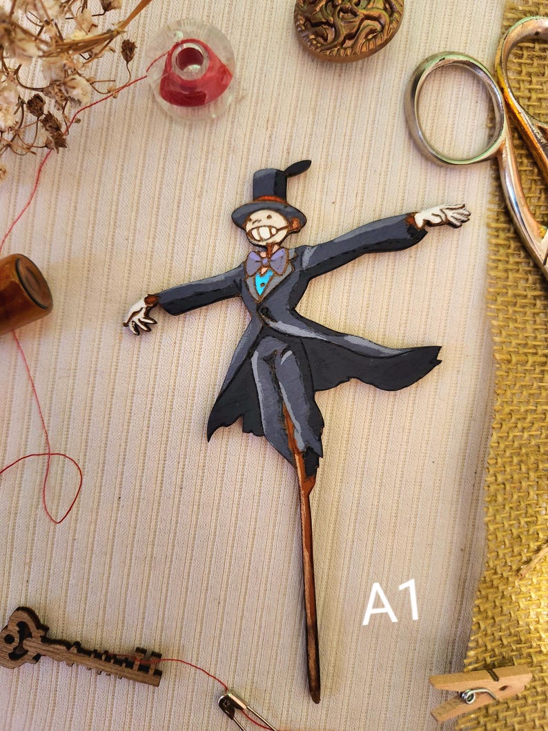 May include: A wooden cutout of a character from the movie Howl's Moving Castle. The character is wearing a black suit, a blue bow tie, and a top hat. The cutout is painted in shades of gray, blue, and black. The character is holding out its arms and has a friendly expression on its face. The cutout is labeled "A1".