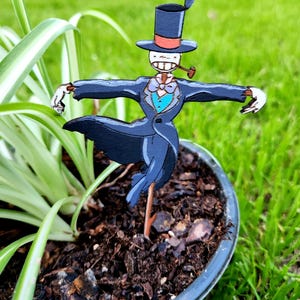 May include: A wooden scarecrow figurine wearing a black suit, a red and white top hat, and a blue bow tie. The scarecrow is holding a pipe in its mouth and is standing in a pot of soil.