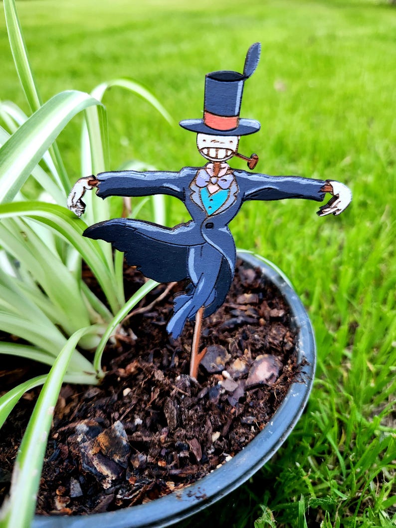 May include: A wooden scarecrow figurine wearing a black suit, a red and white top hat, and a blue bow tie. The scarecrow is holding a pipe in its mouth and is standing in a pot of soil.