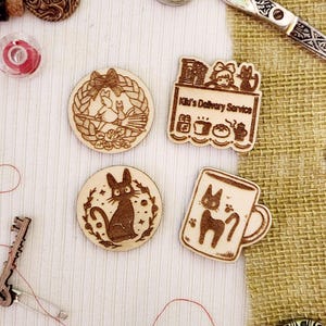 May include: Four wooden laser-cut ornaments featuring characters from the animated film 'Kiki's Delivery Service'. The ornaments depict Kiki on a broomstick, Kiki's cat Jiji, Kiki's delivery service shop, and Jiji in a mug.