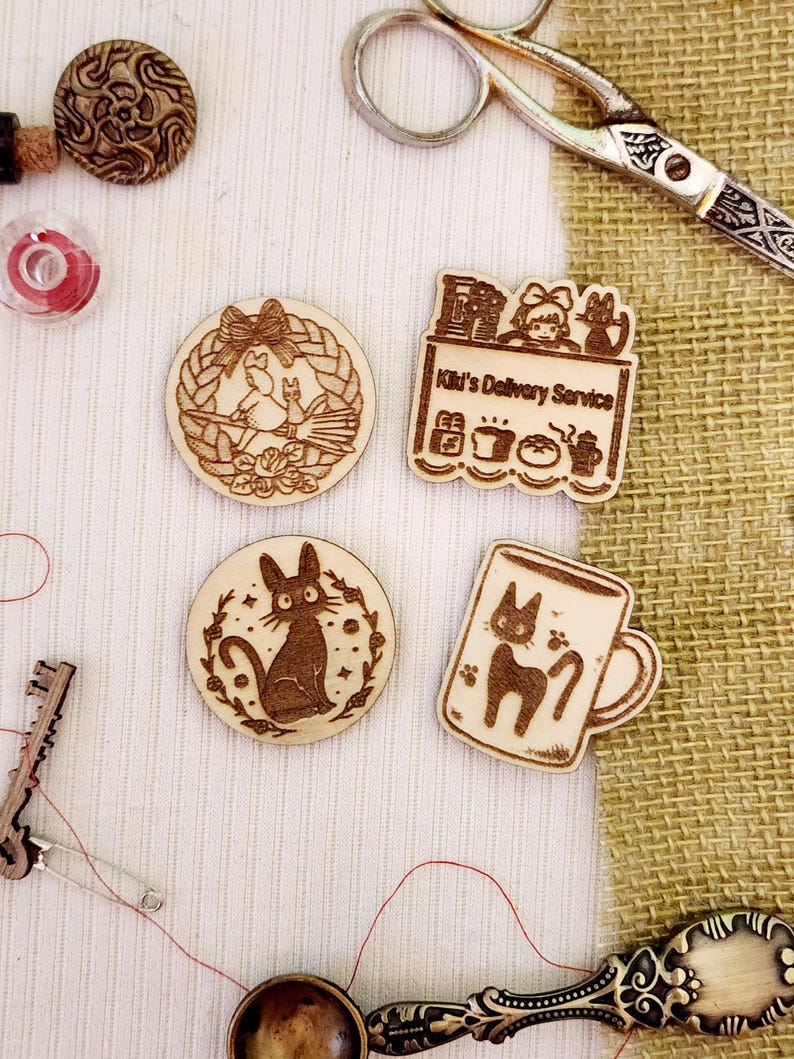 May include: Four wooden laser-cut ornaments featuring characters from the animated film 'Kiki's Delivery Service'. The ornaments depict Kiki on a broomstick, Kiki's cat Jiji, Kiki's delivery service shop, and Jiji in a mug.
