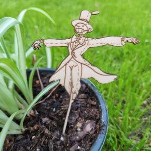 May include: A wooden cutout of a scarecrow wearing a top hat and a tuxedo, standing in a pot of soil. The scarecrow has a long, thin body and is holding its arms out to the sides.