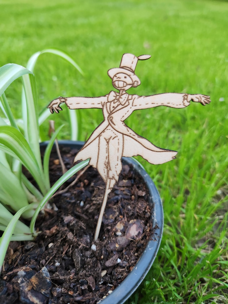 May include: A wooden cutout of a scarecrow wearing a top hat and a tuxedo, standing in a pot of soil. The scarecrow has a long, thin body and is holding its arms out to the sides.