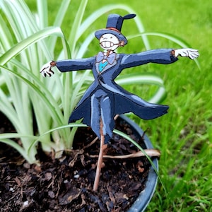 May include: A wooden figurine of a scarecrow wearing a black suit, a top hat, and a bow tie. The scarecrow is standing in a pot with green grass and brown mulch.