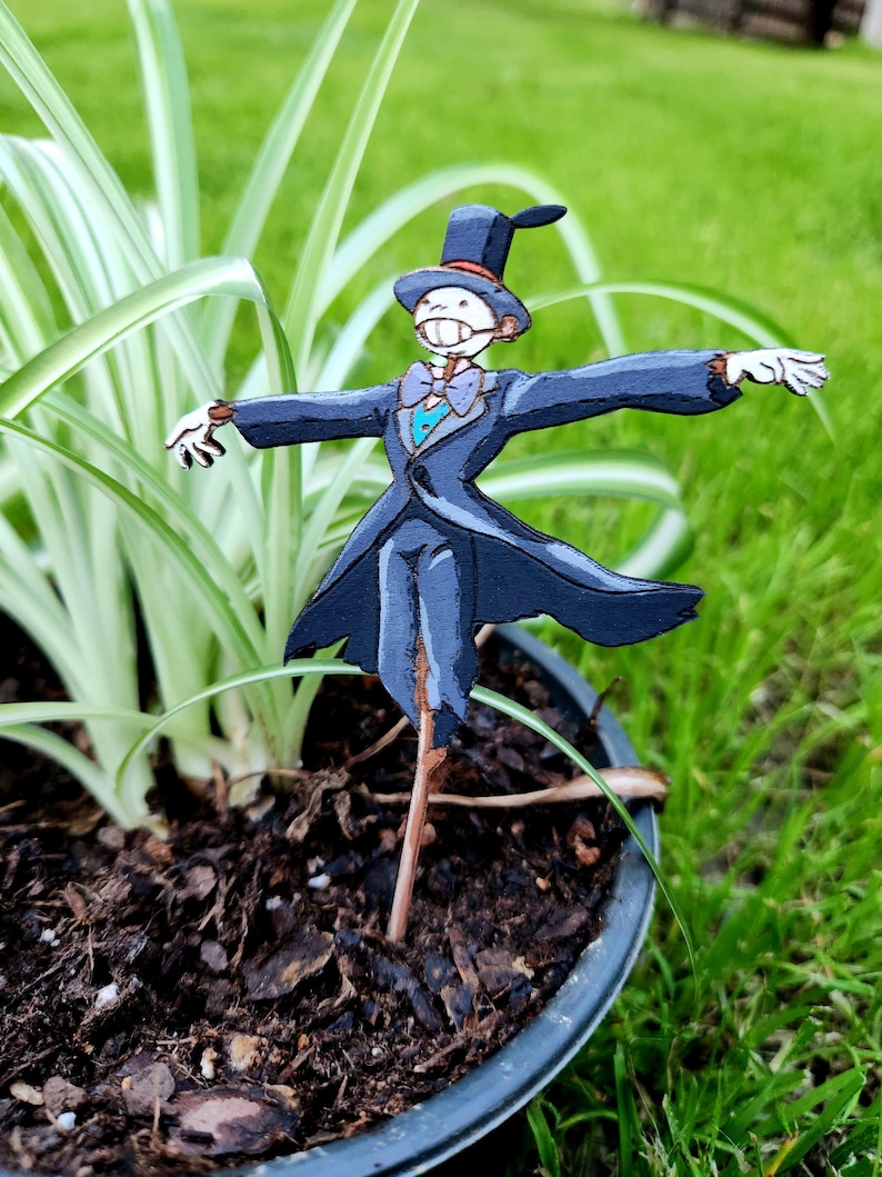 May include: A wooden figurine of a scarecrow wearing a black suit, a top hat, and a bow tie. The scarecrow is standing in a pot with green grass and brown mulch.