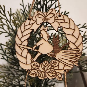 K&ouml;nnte beinhalten: Weihnachtsbaumschmuck aus Holz mit Kiki aus Kikis Lieferdienst, die auf einem Besen mit ihrer schwarzen Katze Jiji fliegt. Der Schmuck ist in Form eines Kranzes mit einer Schleife oben und dem Text "Kiki's Delivery Service" auf einem separaten Anh&auml;nger.