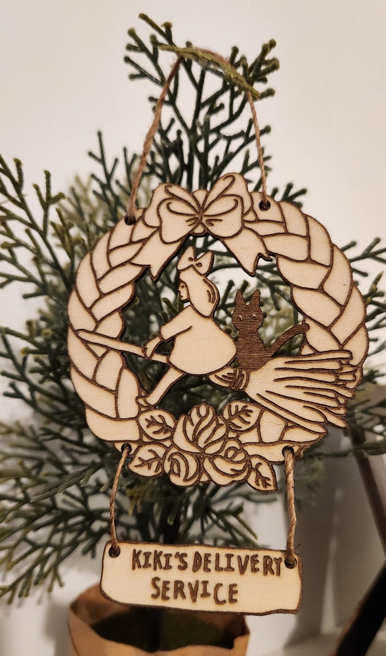 K&ouml;nnte beinhalten: Weihnachtsbaumschmuck aus Holz mit Kiki aus Kikis Lieferdienst, die auf einem Besen mit ihrer schwarzen Katze Jiji fliegt. Der Schmuck ist in Form eines Kranzes mit einer Schleife oben und dem Text "Kiki's Delivery Service" auf einem separaten Anh&auml;nger.