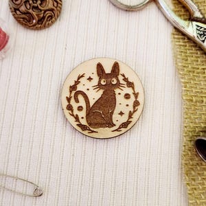 May include: A wooden round pin with a black cat illustration surrounded by a floral wreath. The cat has large eyes and a whimsical expression.