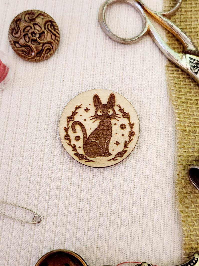 May include: A wooden round pin with a black cat illustration surrounded by a floral wreath. The cat has large eyes and a whimsical expression.