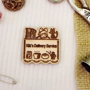 May include: A wooden cutout of Kiki from the movie Kiki's Delivery Service. The cutout features Kiki with her cat Jiji, a delivery cart, and a variety of items including a loaf of bread, a cup of coffee, and a teapot. The text "Kiki's Delivery Service" is written on the cart.