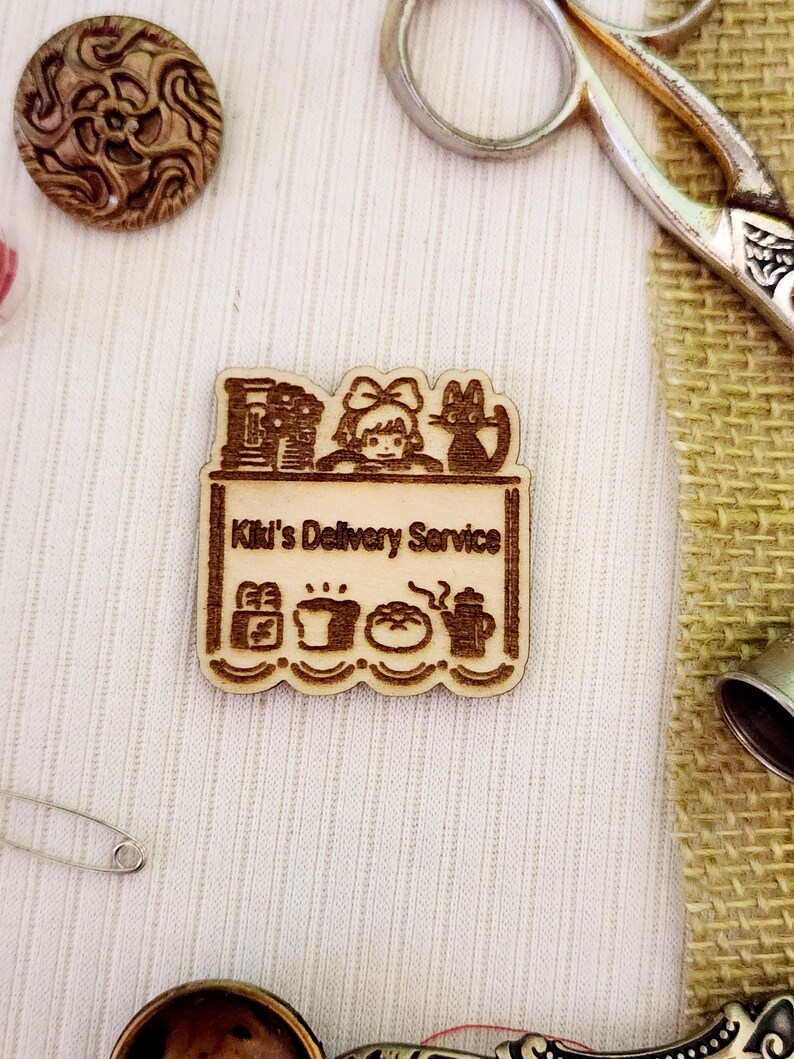 May include: A wooden cutout of Kiki from the movie Kiki's Delivery Service. The cutout features Kiki with her cat Jiji, a delivery cart, and a variety of items including a loaf of bread, a cup of coffee, and a teapot. The text "Kiki's Delivery Service" is written on the cart.