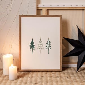 May include: A framed print featuring three stylized green Christmas trees with star accents. The artwork is set against a cream background within a light brown wooden frame. Two white candles and a black star add to the holiday theme.