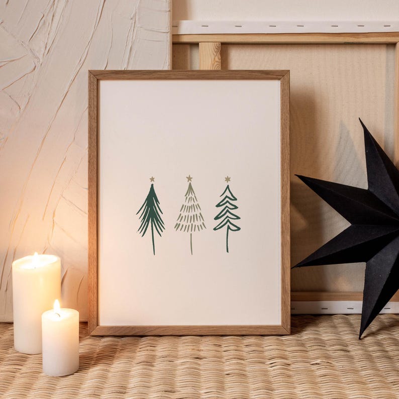 May include: A framed print featuring three stylized green Christmas trees with star accents. The artwork is set against a cream background within a light brown wooden frame. Two white candles and a black star add to the holiday theme.
