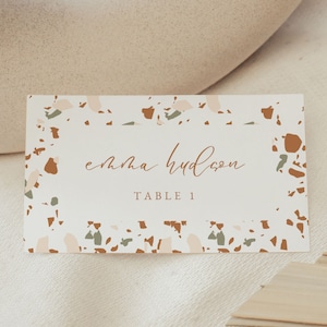 May include: A white place card with a terrazzo pattern border. The text "emma hudson" is written in brown cursive script. The text "TABLE 1" is written in brown sans-serif font.