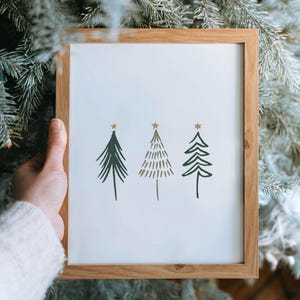 May include: A framed print featuring three stylized Christmas trees in shades of green and gold, each topped with a gold star. The trees are set against a white background within a natural wood frame, perfect for holiday decor.